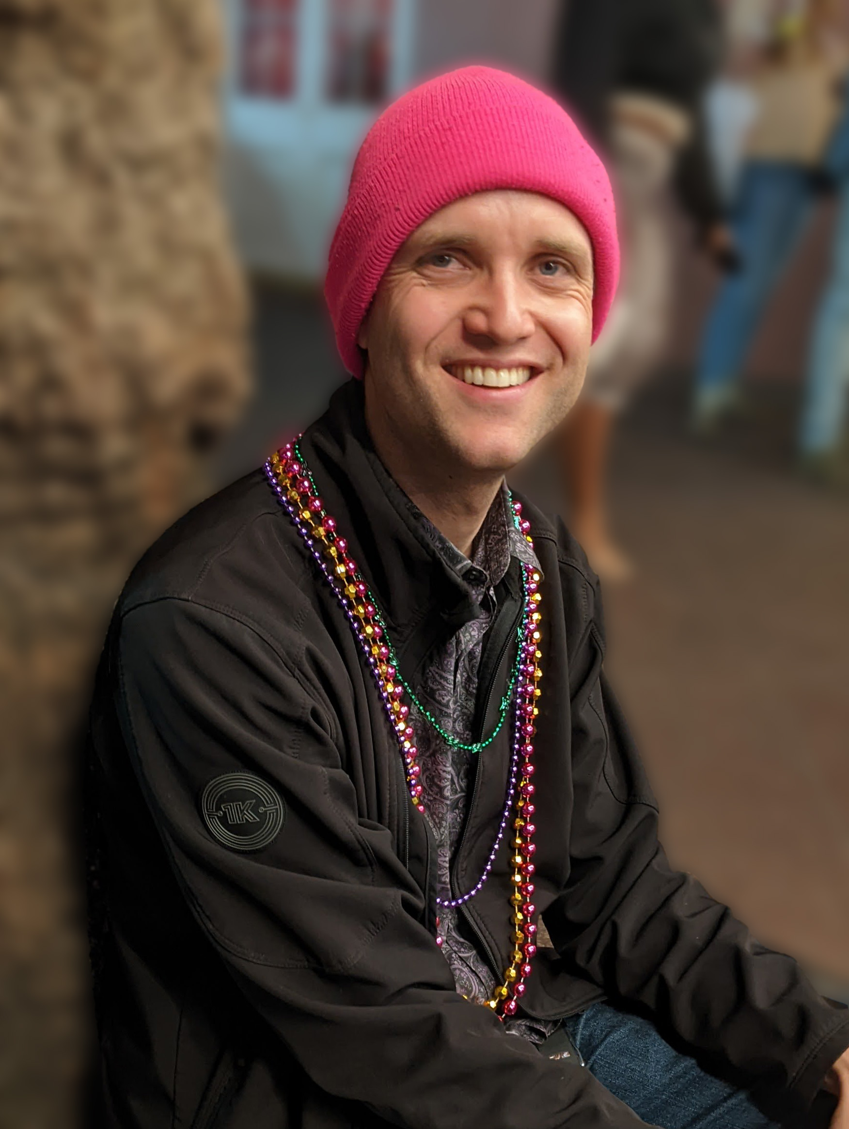 Cody Harris: a man in his 30s wearing a black jacket, pink beanie, and Mardi Gras beads, leaning against a tree. The background is blurred.